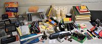 Wide view of assorted office supplies arranged on a table including staplers, calculators, pens, paper clips, and books.