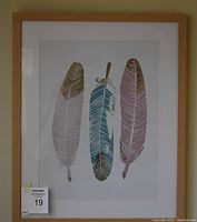 Framed print featuring three stylized feathers in pink, blue, and gold accents within a light wooden frame, shown from a frontal view.
