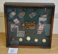 Framed golf memorabilia with four vintage club heads, brass golf emblem, five golf balls, pins, and golf paper clippings on green felt background.