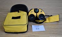 Binoculars with compass and adjustable eyecups next to matching yellow and black carrying bag on wooden table
