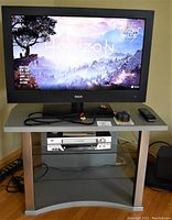 RCA 32 inch flat screen TV powered on displaying game screen, placed on TV stand with glass shelves. JVC VHS player present on lower shelf with remote and cables on top.