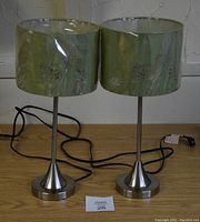 Full view of the two matching green table lamps side-by-side on a table, showing metallic bases and green floral cutout patterned shades with cords attached.