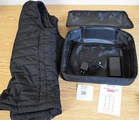 Photo of the black heated vest laid out beside the open black zippered carrying case containing charger and battery pack, plus instruction card and small info card with size 36.