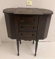 Front view of antique Martha Washington sewing table showing closed side compartments, three front drawers with wooden knobs, and four turned legs.