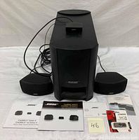 Bose Acoustimass module with two satellite speakers, interface module, and manuals arranged on surface.