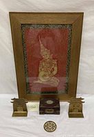 Framed Buddha portrait, Chinese vanity box, pair of brass bookends, and brass emblem arranged on a white cloth background.