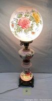 Full view of the vintage lamp showing the hand-painted floral glass globe shade with pink and yellow roses and the pink floral glass base, brass accents, and winding key.