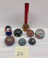 Image of eight Murano glass paperweights arranged in front of a white wall, varying sizes and colorful millefiori designs, plus a tall red Murano glass bud vase with a round clear base containing an internal flower design.