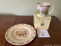 Plate and mug with original box on wooden surface, showing full items in lot