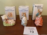 Three Beatrix Potter figurines with boxes behind them on wooden table. Left figurine is lying rabbit with bonnet, center is standing rabbit holding basket, right is rabbit in red coat. Boxes labelled Royal Albert and Beswick are present.