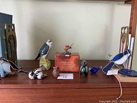 Front view of bird figurine collection including the Merlin Scotch Whiskey bottle in original orange box and various glass and ceramic bird figurines arranged on a wooden surface.