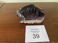 Front view of the purple amethyst glass water buffalo figurine placed on a wooden table next to a maxsold number card.