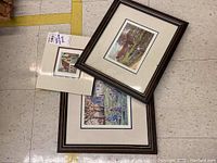 Three framed prints arranged on floor: the smallest is a scene of a historic red-roofed building, one larger depicts outdoor scene with children, third depicts a fishing scene with several people near water