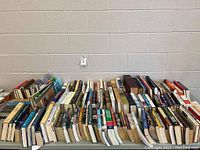 Photo showing multiple rows of hardcover and paperback books arranged on a table against a wall, titles mostly obscured but various genres evident