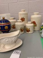 Full view of five ceramic cannisters with fruit designs and a white ceramic gravy boat with matching tray on a green table.