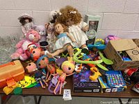 Full view of assortment on table showing dolls, plush toys, furniture, puzzle, tackle box, and mugs