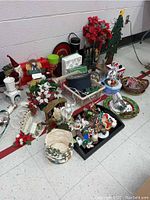 Photo shows an assortment of Christmas decorations including a wood tree vase with red faux flowers, trays, mugs, tins, ornaments, baskets, a Santa figurine, and a snow globe.