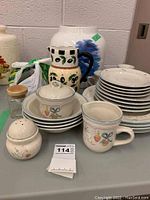 Photo of various dishes including stacked plates, bowls, a covered dish, and pitchers with painted fruit and leaf designs.