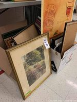 Photo showing multiple framed pictures stacked and two cardboard boxes with a large Italian double-sided poster against the wall.