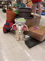 Wide view showing plastic flower pots in different sizes and colors, the elephant planter, oasis foam, and some wooden shelves in background