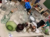Various glassware including clear glass bottles, dark brown glass jug, green glass bowl, cream-colored jug, and ceramic dishes on a table.