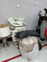 Photo showing vintage white hamper with floral bedspread and assorted throw pillows stacked on and around it next to a table lamp