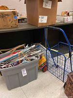 Grey bin filled with assorted hangers and wire racks next to the blue metal shopping cart and a multicolored mini ironing board.