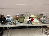 Wide view of entire lot of kitchen accessories including crock pots, stoneware bowls, glassware, and small appliances on table against a wall