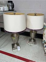 Pair of matching lamps with urn-shaped metal bases and white lampshades trimmed with purple beads, placed on a floor near wall with a floral cushion nearby.