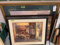 Three large framed prints leaning against a wall, visible details of one print showing antique shop scene.