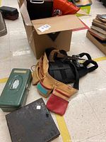 Wide shot showing green metal toolbox, tool belt with red and turquoise pouches, black back brace, and cardboard box in background.