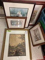 Five framed prints stacked and leaning against a wall, showing varying winter and country scenes including a schoolhouse.