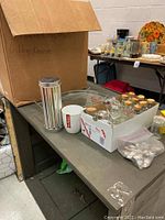 Shoebox containing multiple glass spice bottles and jars, most with lids, with additional kitchen items on table including white mug with red logo, plastic containers, and metal rack.