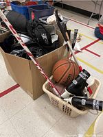Photo of sports equipment including basketballs, badminton rackets, hockey stick, Bauer hockey skates, and a cardboard box with gear inside.