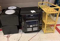 Photo of RCA stereo system with two black speakers and yellow woven wicker shelf on floor.