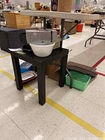 Black square occasional table with a white mixing bowl on top, boxes, and green and red dishes under and around the table.