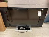Photo showing the entire Prima 32 inch TV on a stand with remote control placed on base.