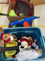 Photo showing blue toy guns, stuffed deer toy in holiday costume, a blonde doll, and various toys in a blue plastic bin and cardboard boxes.