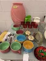 Photo showing large coral vase, pastel colored jumbo coffee mugs, red-topped goblets, handmade pitcher, and jar on the table.