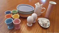 Photo of six pastel-colored egg cups, matching plates, white china creamer holding white egg spoons, two white salt/pepper shakers, and a tall pink vase/container with gold rim.