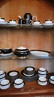 Full view of the display cabinet showing various plates, cups, coffee pot, serveware items all in black, white and gold Fitz and Floyd Renaissance pattern.