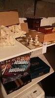 Shelf with two chess sets including a boxed set with green and cream pieces and a marble-like carved set with packaging paper beside it, plus other wooden boxes for games.