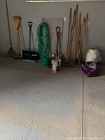 Photo showing assortment of gardening tools, garden hose, watering can, booster cables and various containers on garage floor.
