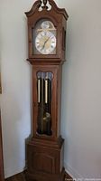 Full front view of grandmother clock showing wooden case, hood, finial, glass door, pendulum, weights, and base cabinet