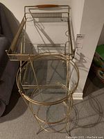 Full front and top view of the brass and glass bar cart showing handle, two glass shelves, perforated brass sides, and wheels.