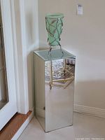 Full view of the green glass vase with silver metal leaf vine stand atop the mirrored pedestal against a light wall.