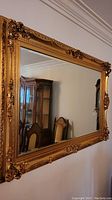 Full frontal photo of large ornate wall mirror with bronze/gold resin frame and beveled glass, mounted on wall with dining room chairs reflected