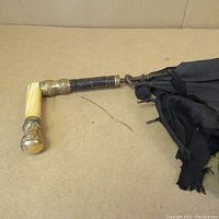 Side view of the antique parasol showing the carved bone handle with brass fittings attached to black fabric folded and in tattered condition.