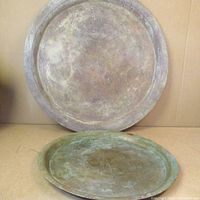 Two round trays displayed upright against a wall, one larger copper tray on the back and a smaller brass tray in front.