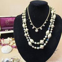 Pearl and crystal bead necklaces displayed on a black bust with additional beads and earrings in the background